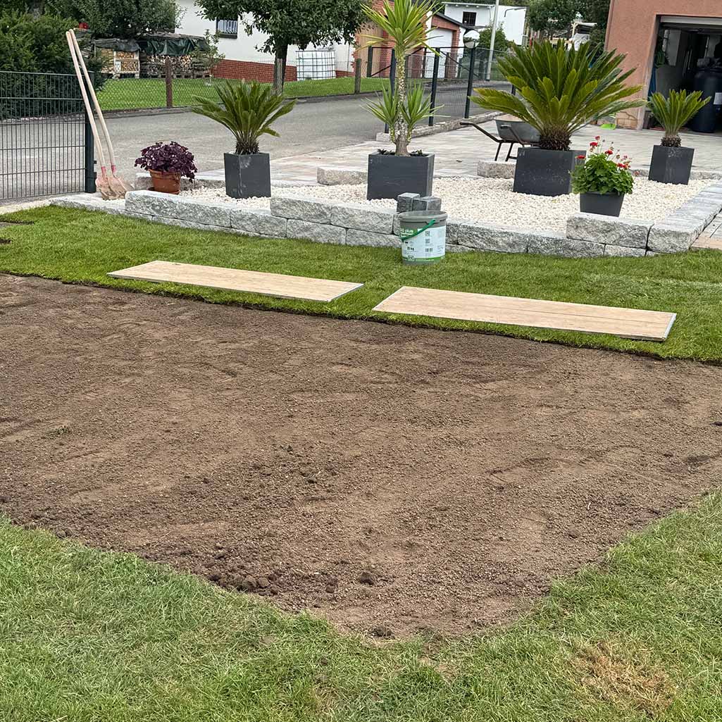 Ein gepflegter Rasen, zwei Bretter, Gartenwerkzeuge und Blumen in Pflanzquartieren, im Hintergrund.