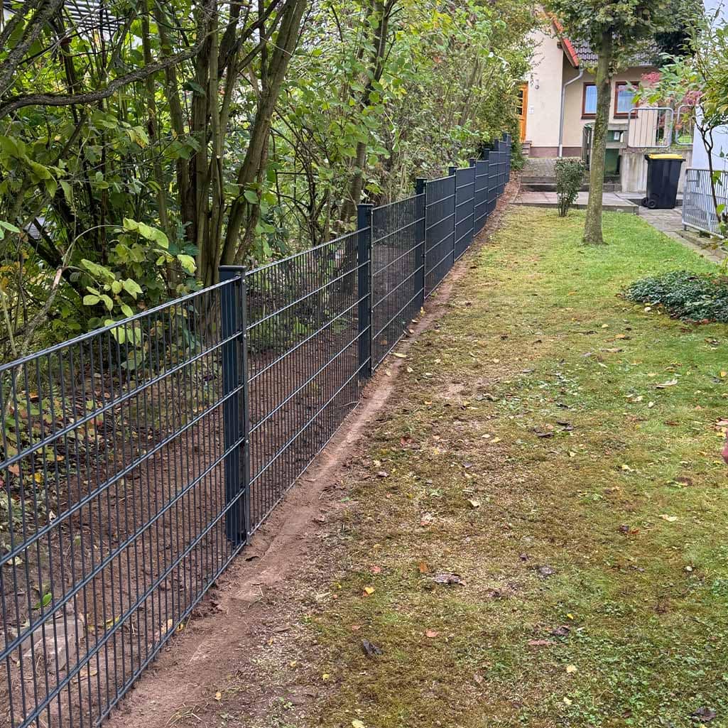 Neuer schwarzer Zaun entlang eines Gartens mit Bäumen und grünem Rasen, Haus im Hintergrund.